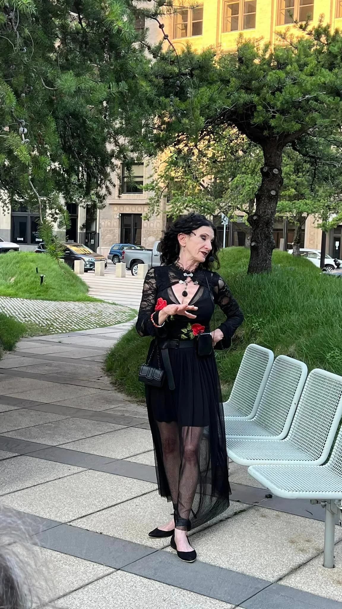 A woman in a black dress is standing next to a bench in a park.