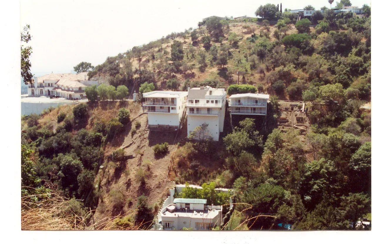 Three white buildings perched on a hillside covered in green trees and shrubs.