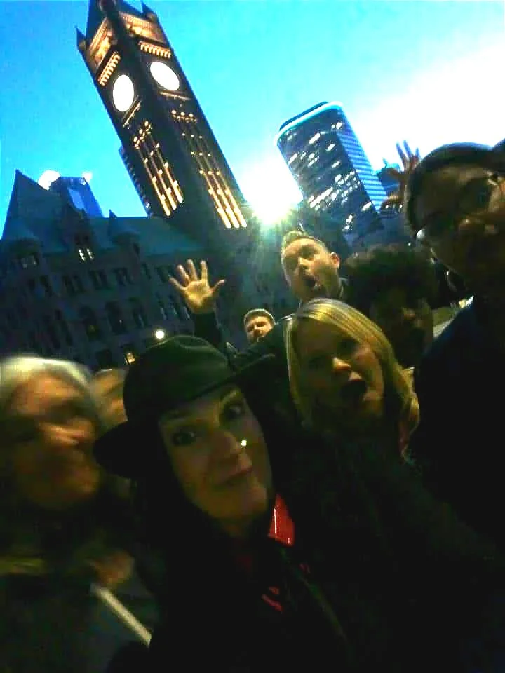 People taking a selfie near a large clock tower at dusk. Some people are smiling, and one is waving.