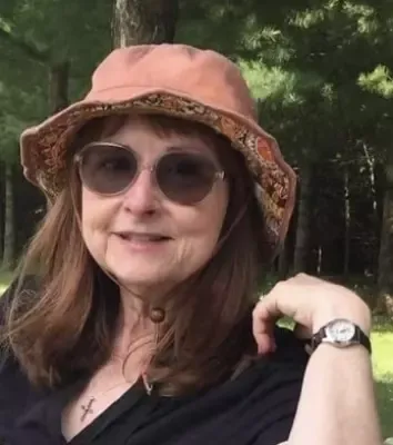 A woman wearing a hat and sunglasses is standing in the woods.