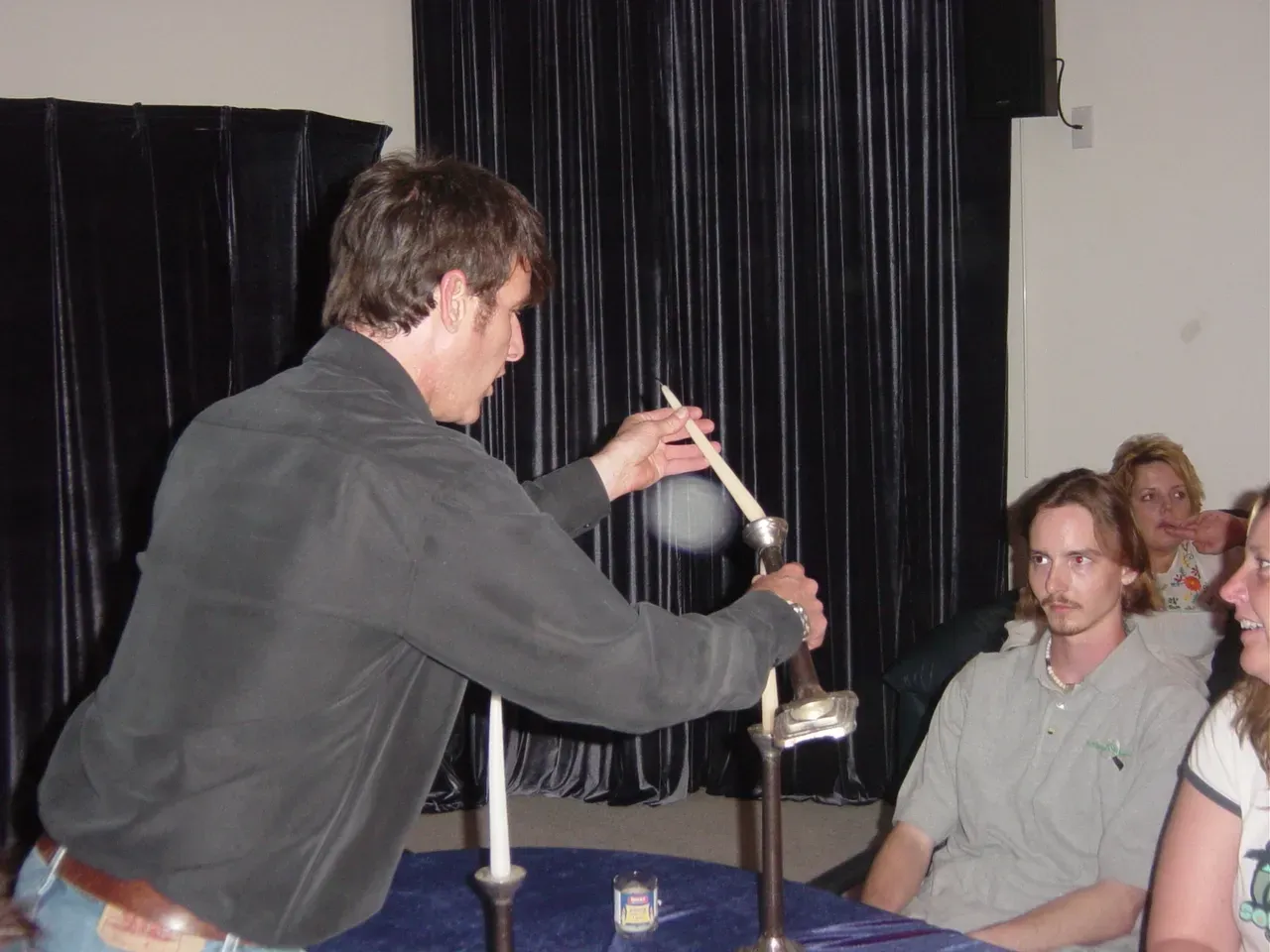 Man in black shirt performing a demonstration with smoke, candles, and an audience.