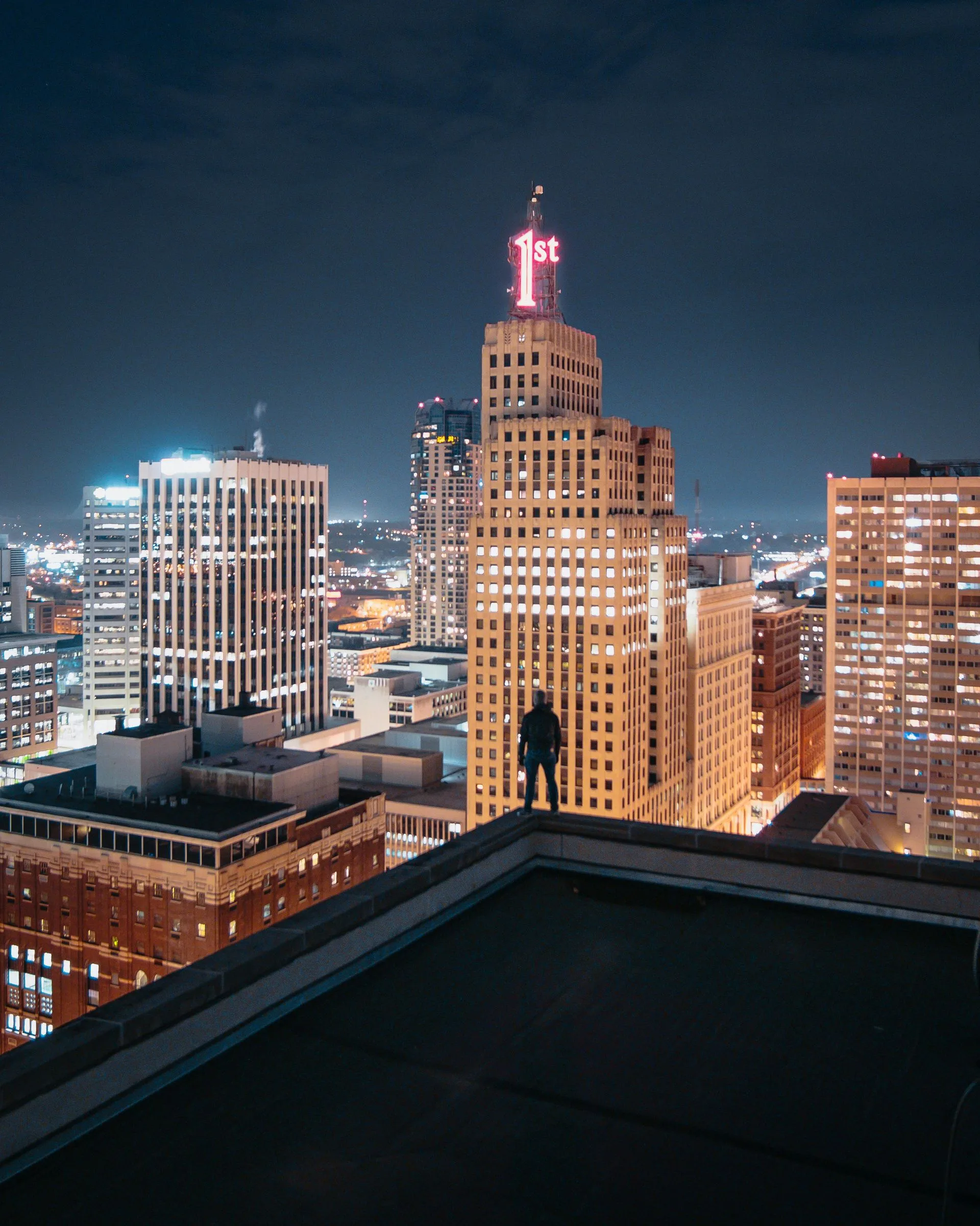 Person on rooftop overlooking city at night, illuminated skyscrapers with neon sign.