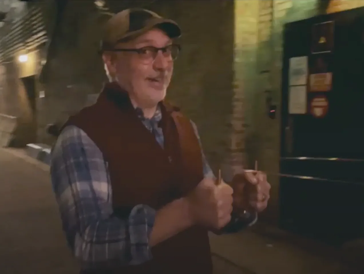 A man wearing glasses and a hat is standing in a dark alleyway.